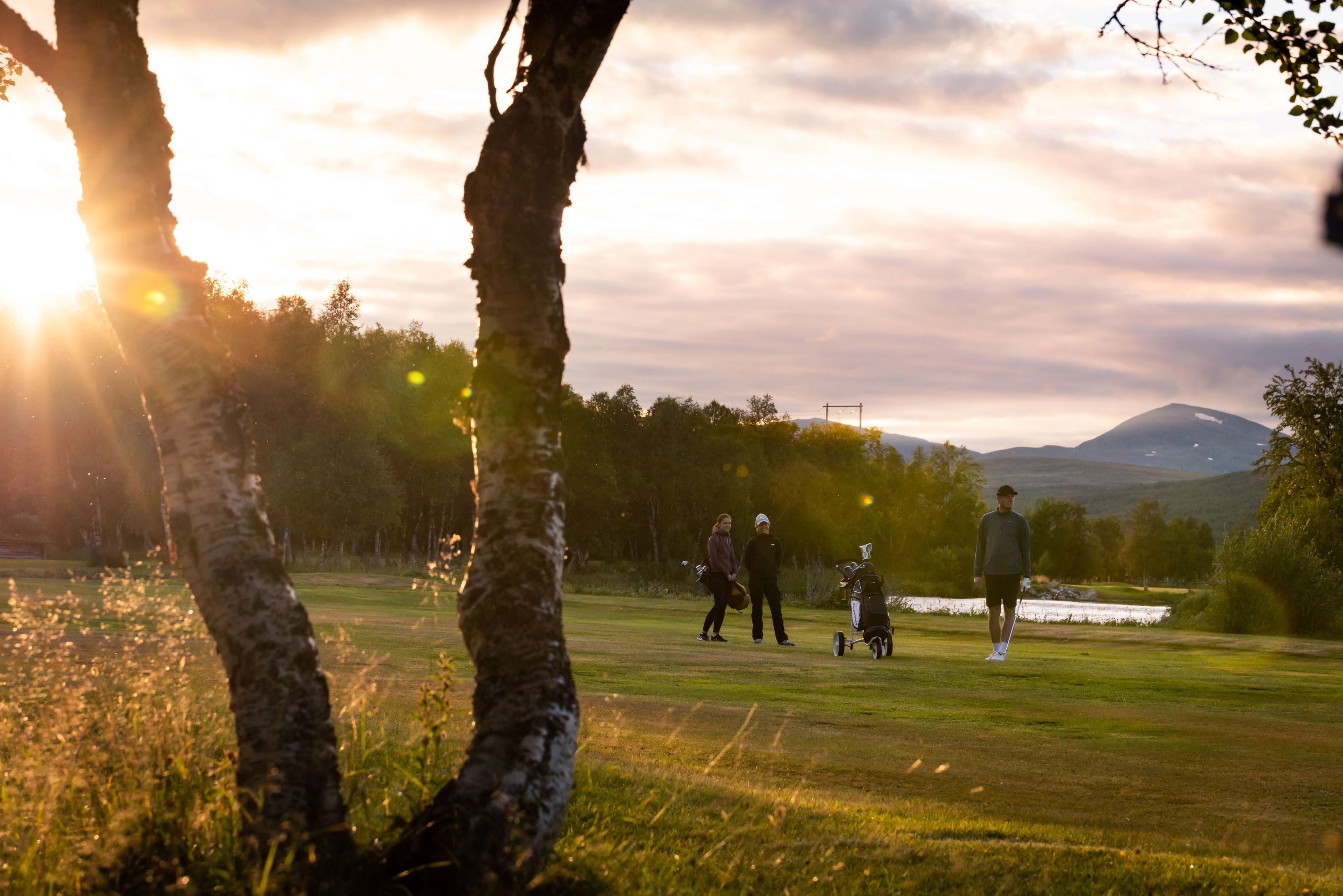 A golf course at sunset with three people, a golf cart, mountains, and tree trunks in the foreground.