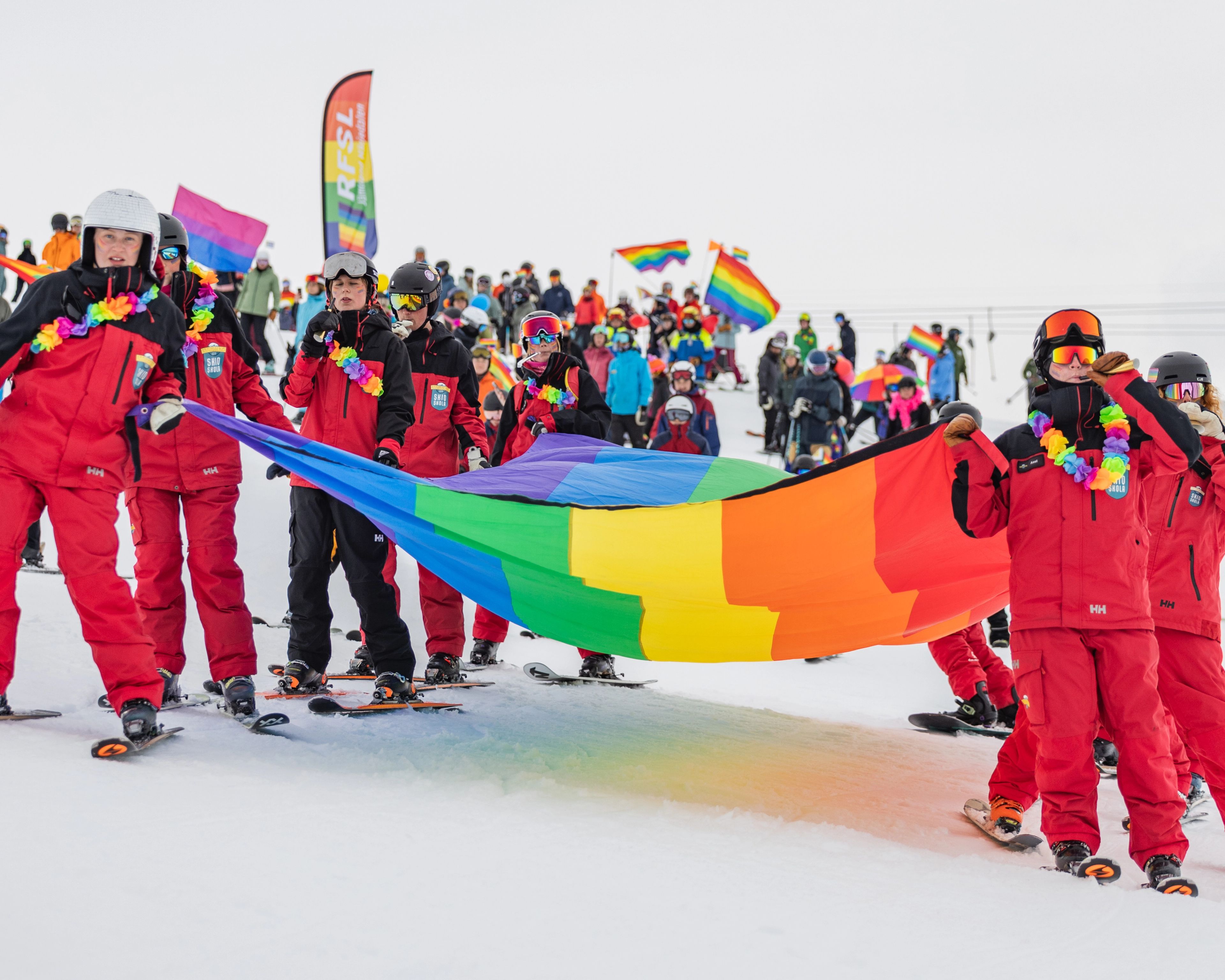 Skiers in red outfits carry a large rainbow Pride flag on a snowy mountain, with more people and flags in the background.