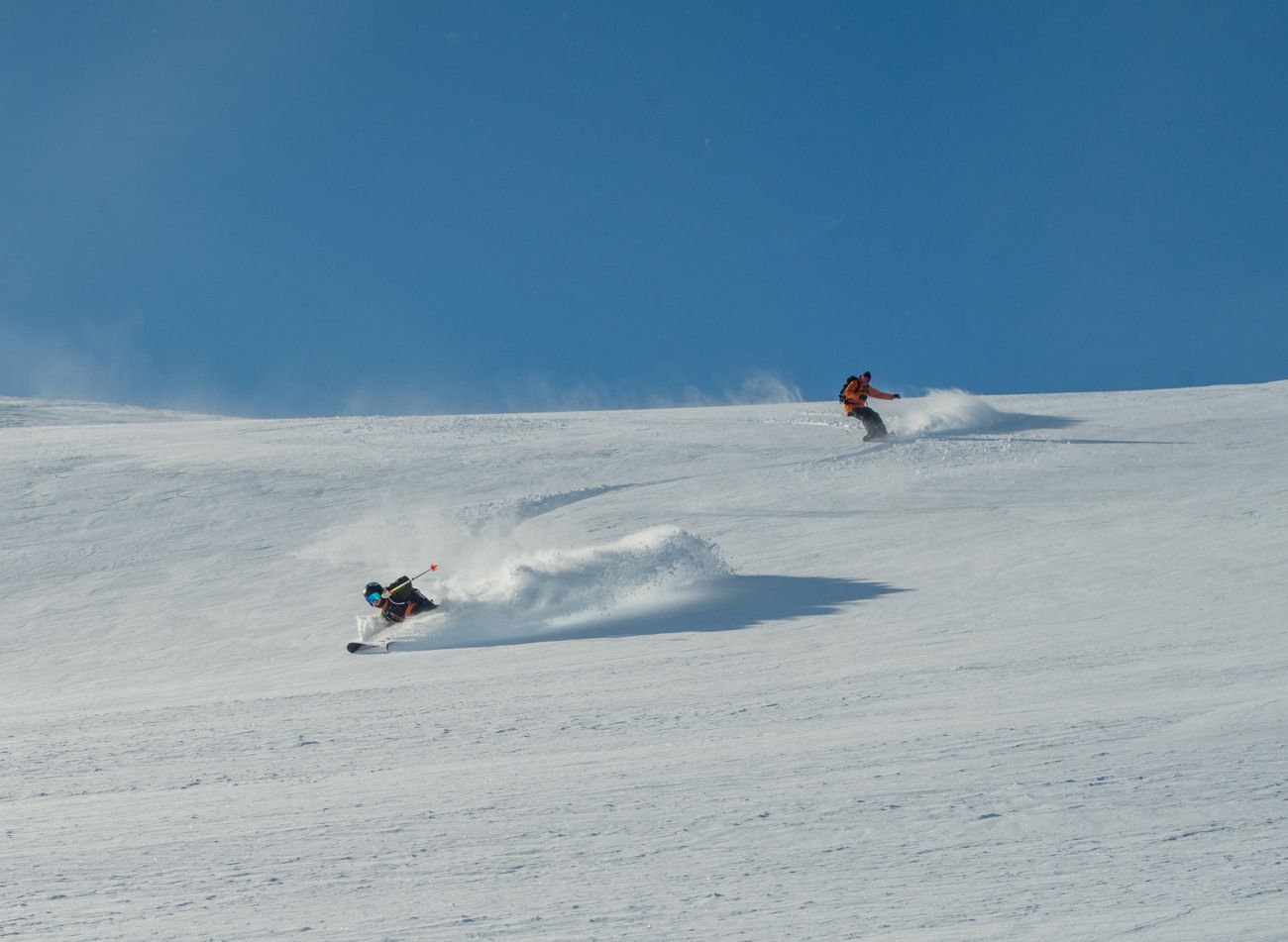 Två personer som åker skidor i lössnö nedför ett snöklätt berg.