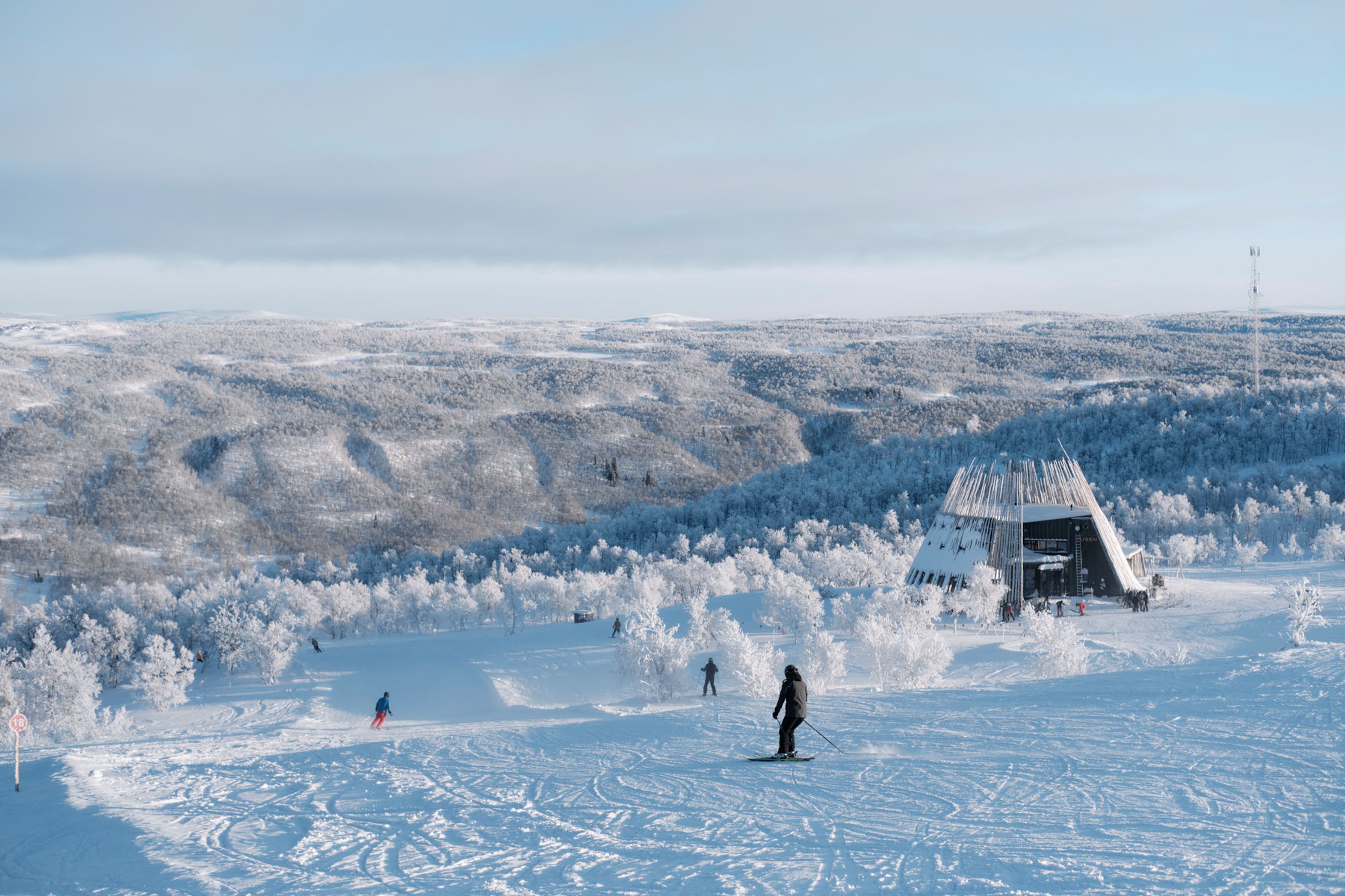 En grupp människor som åker skidor på toppen av en snötäckt backe med restaurang Tusen i bakgrunden.