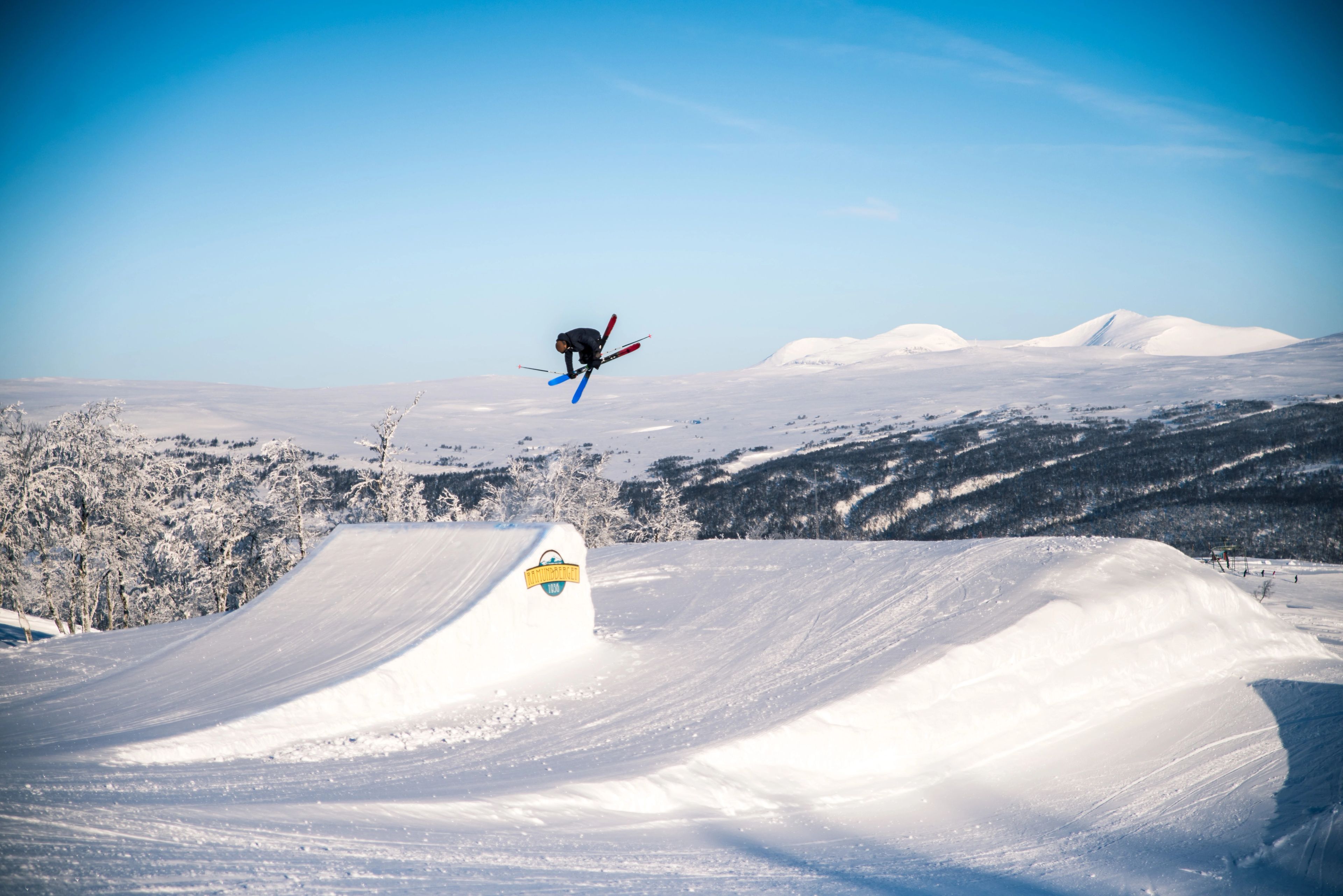 A skier performs an aerial grab trick over a snow jump in a sunny, snowy mountain landscape.