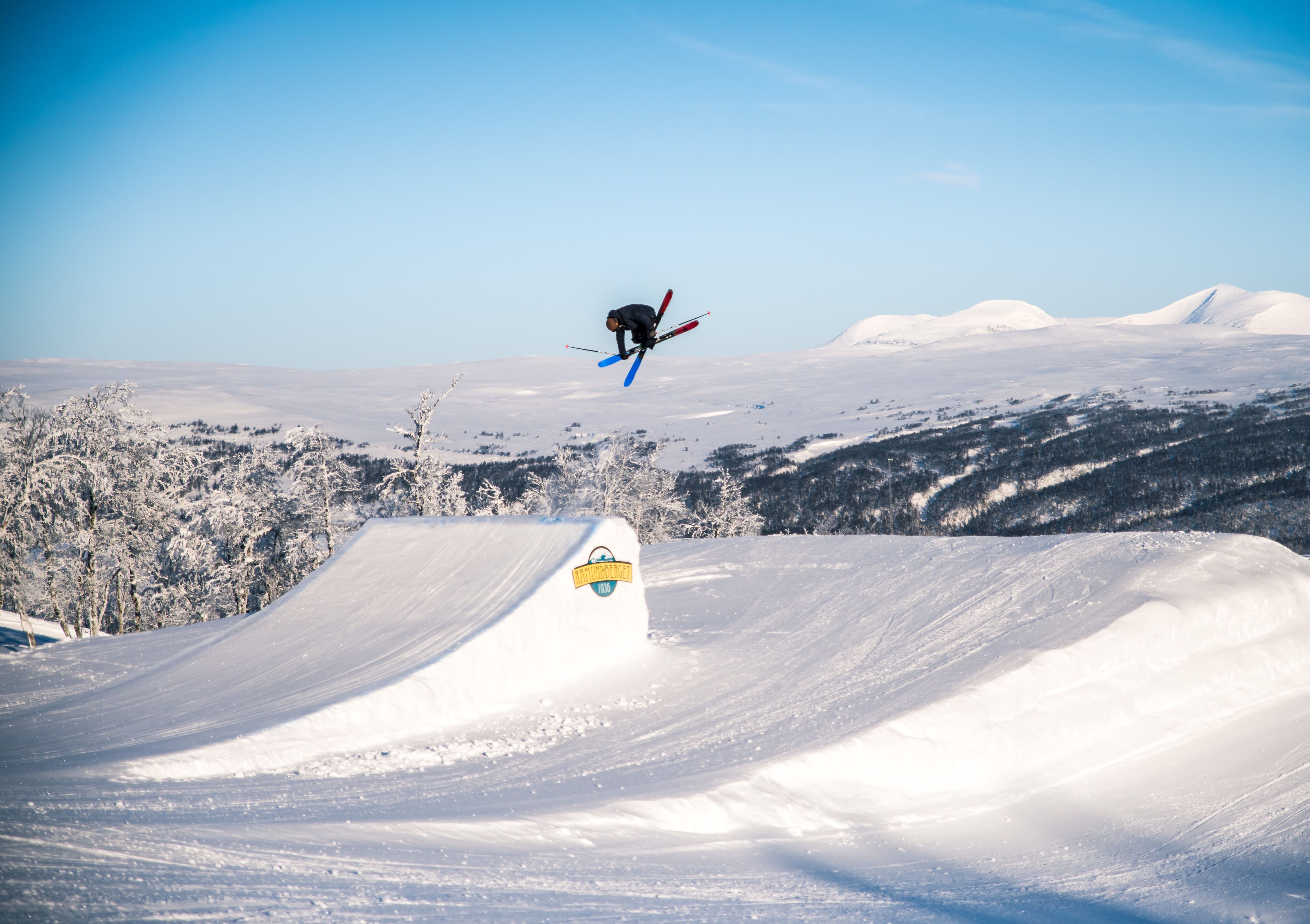 A skier jumps mid-air over a snow ramp in a snowy mountain landscape.