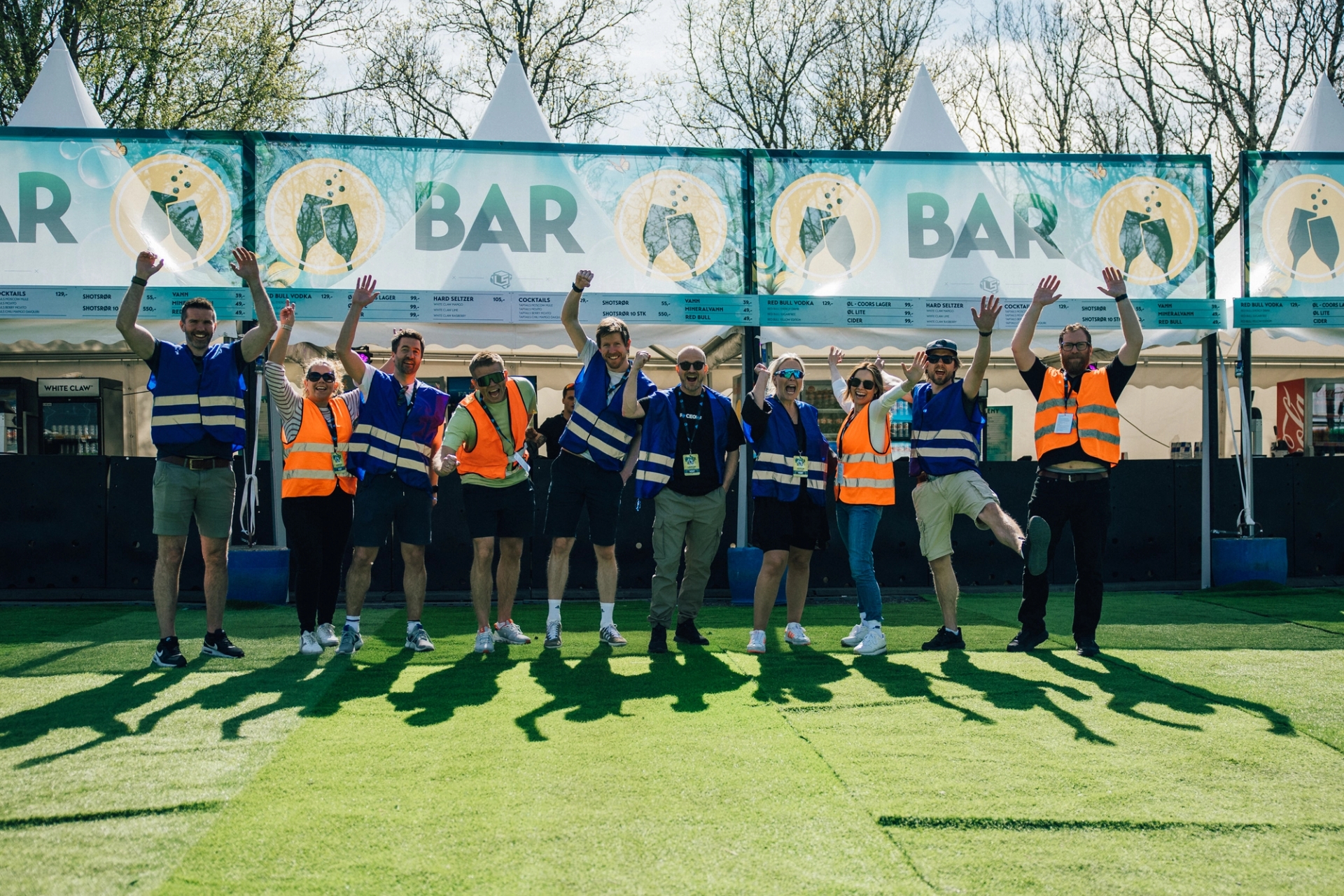 Glade ansatte i oransje og blå sikkerhetsvester feirer foran en festivalbar.