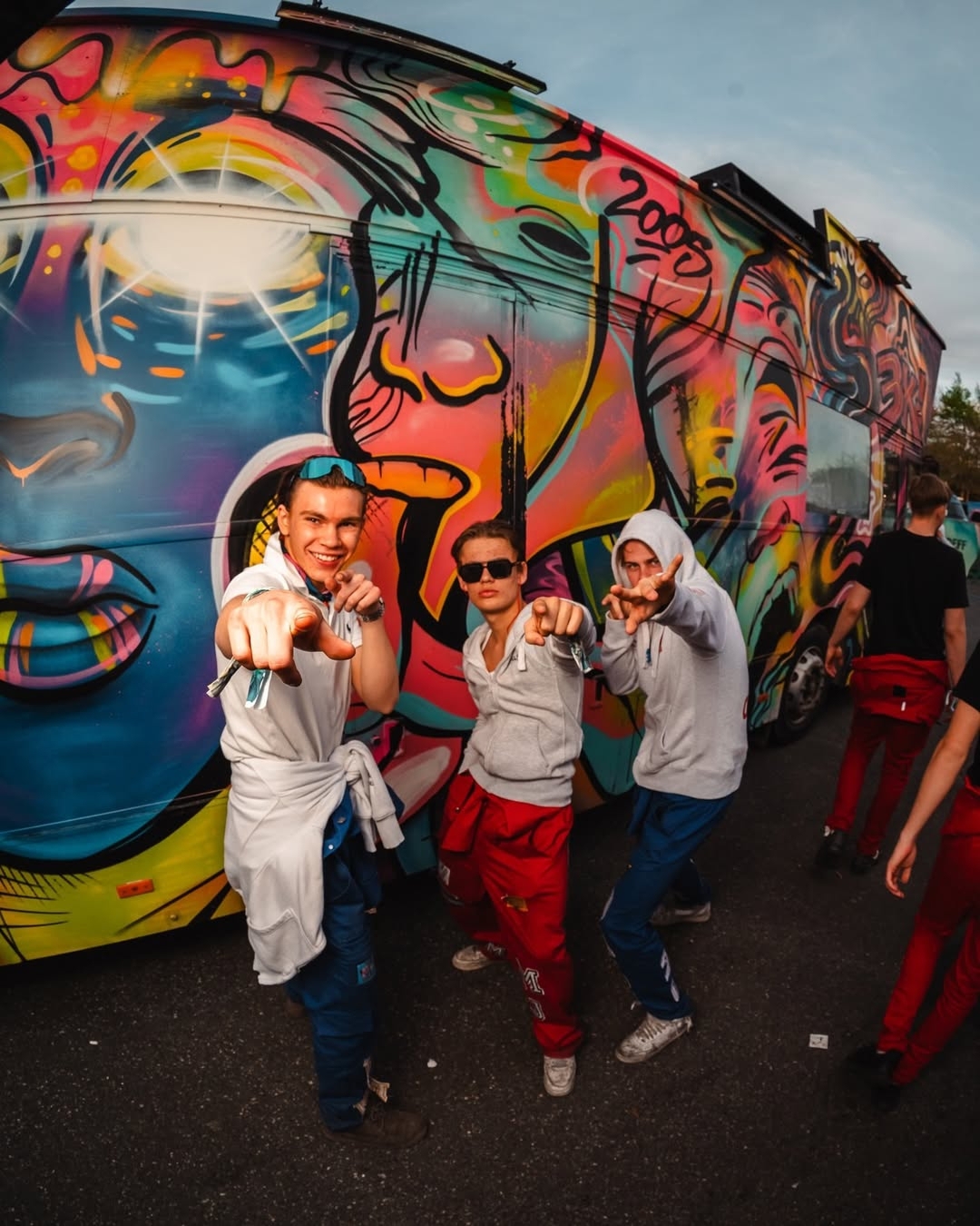 En guttegjeng står foran en fargerik russebuss og peker mot kameraet.