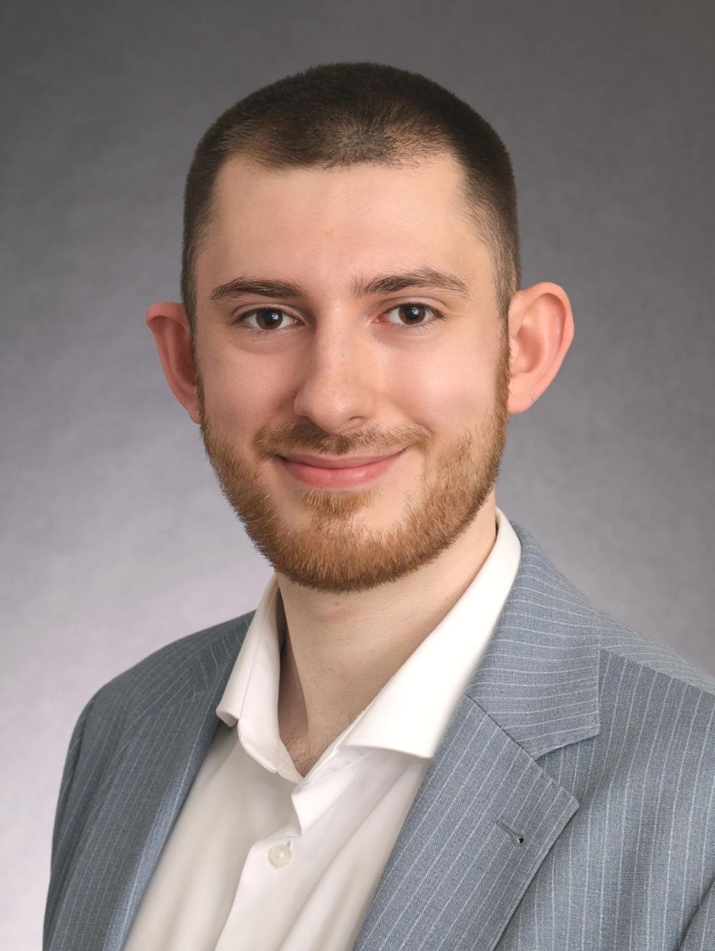 Profilbild von Lino Dotsikas, Jura-Dozent, in grauem Anzug und weißem Hemd vor neutralem Hintergrund.