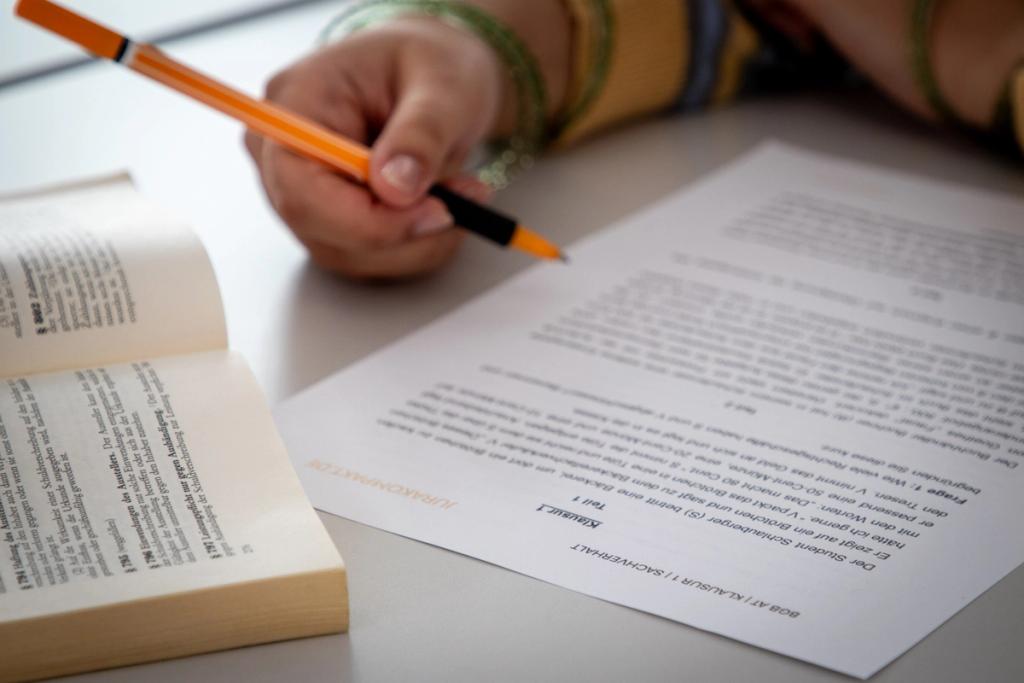 Hand mit orangenem Stift bearbeitet Jurakompakt-Aufgabenblatt neben aufgeschlagenem Gesetzbuch