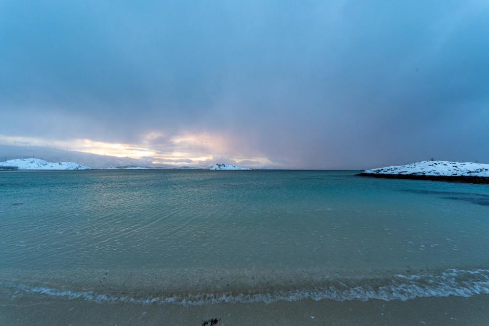 Leuchtend türkisfarbenes Meer läuft über den hellen Strand von Sommarøy aus, dahinter schneebedeckte Gipfel im rosa Licht.