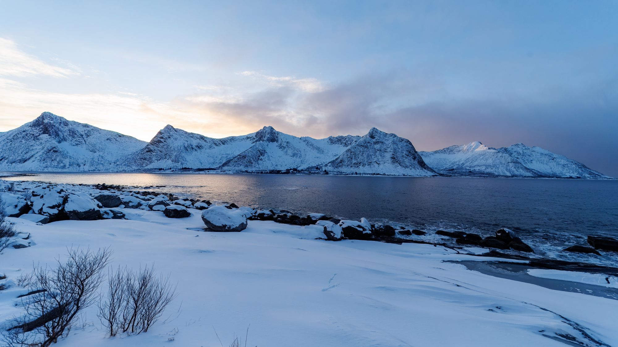 Eine ruhige, verschneite Bucht öffnet den Blick auf die gezackte Felsreihe der Okshornan unter blassem Winterhimmel.