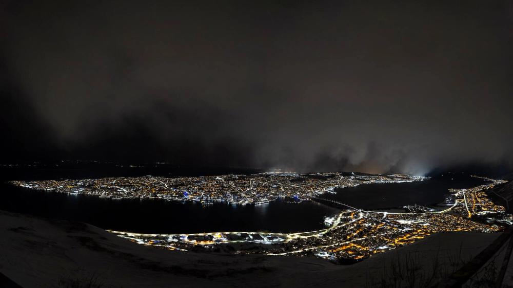 Panoramablick vom Aussichtspunkt Fjellheisen auf das nächtliche Tromsø mit beleuchteter Innenstadt, Brücke und umliegenden Bergen, im Vordergrund Schnee.