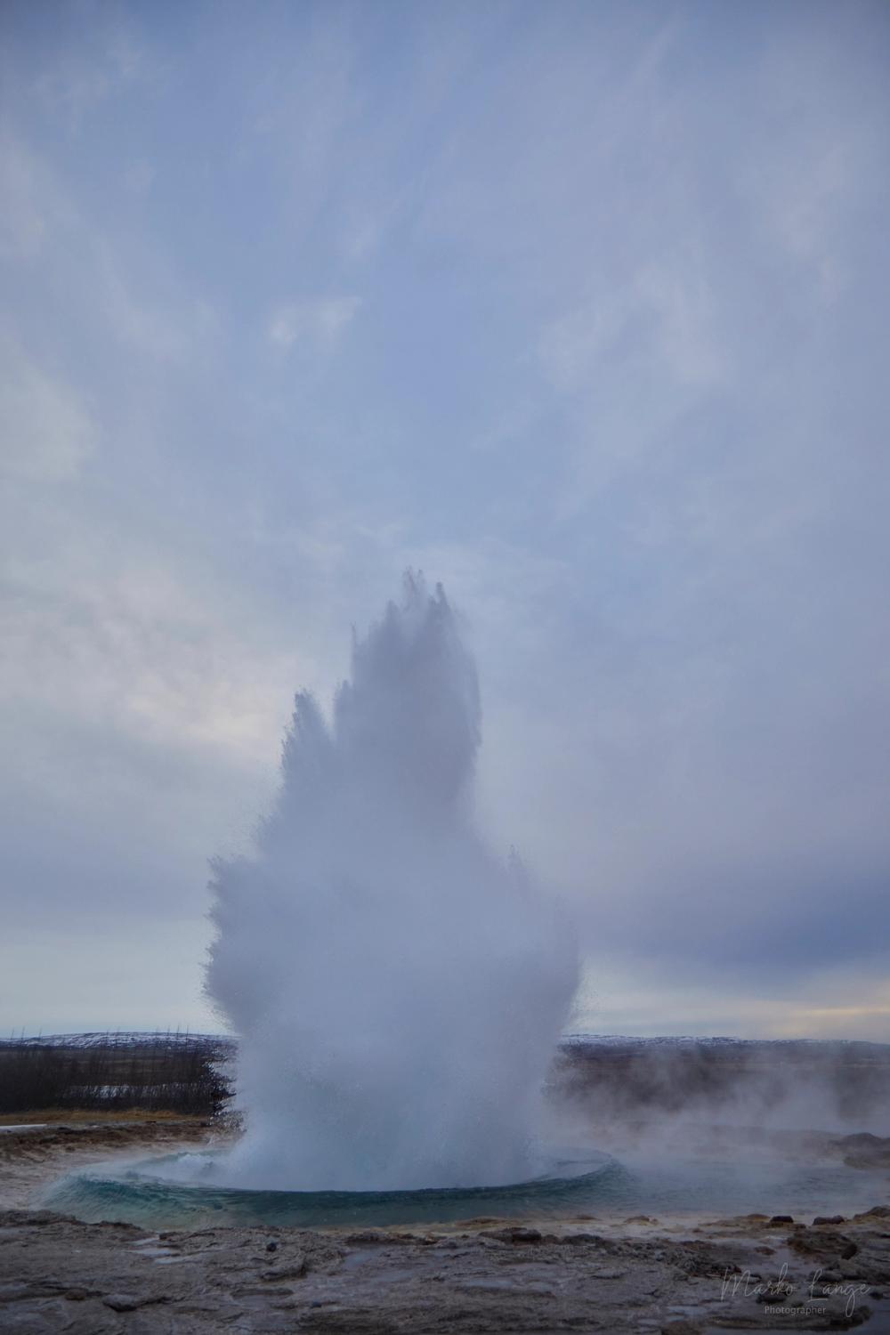 Der ausbrechende Strokkur wirft eine schlanke Fontäne aus Wasser und Dampf senkrecht empor.
