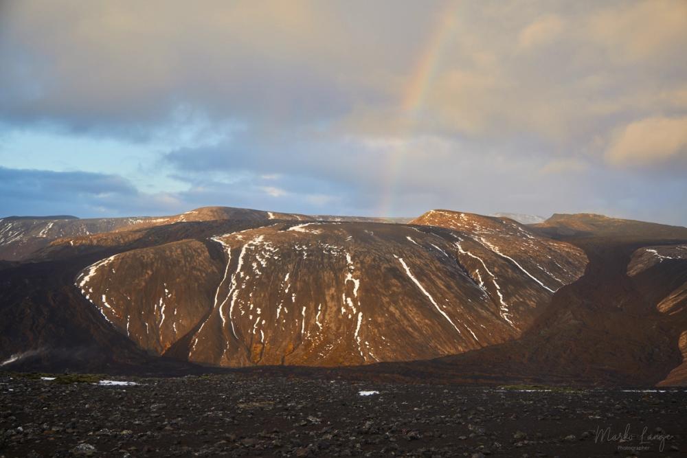 Ein Regenbogen steht über einem sonnenbeschienenen Bergrücken, davor schwarze Lavafelder.