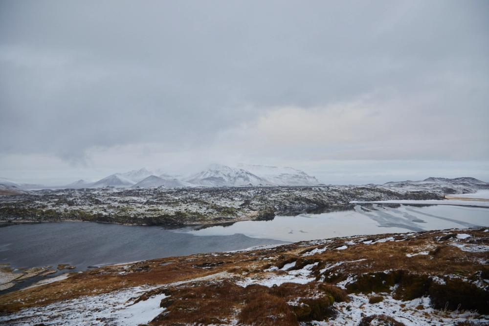 Blick über verschneite Lavafelder auf einen halb zugefrorenen See und schneebedeckte Berge.