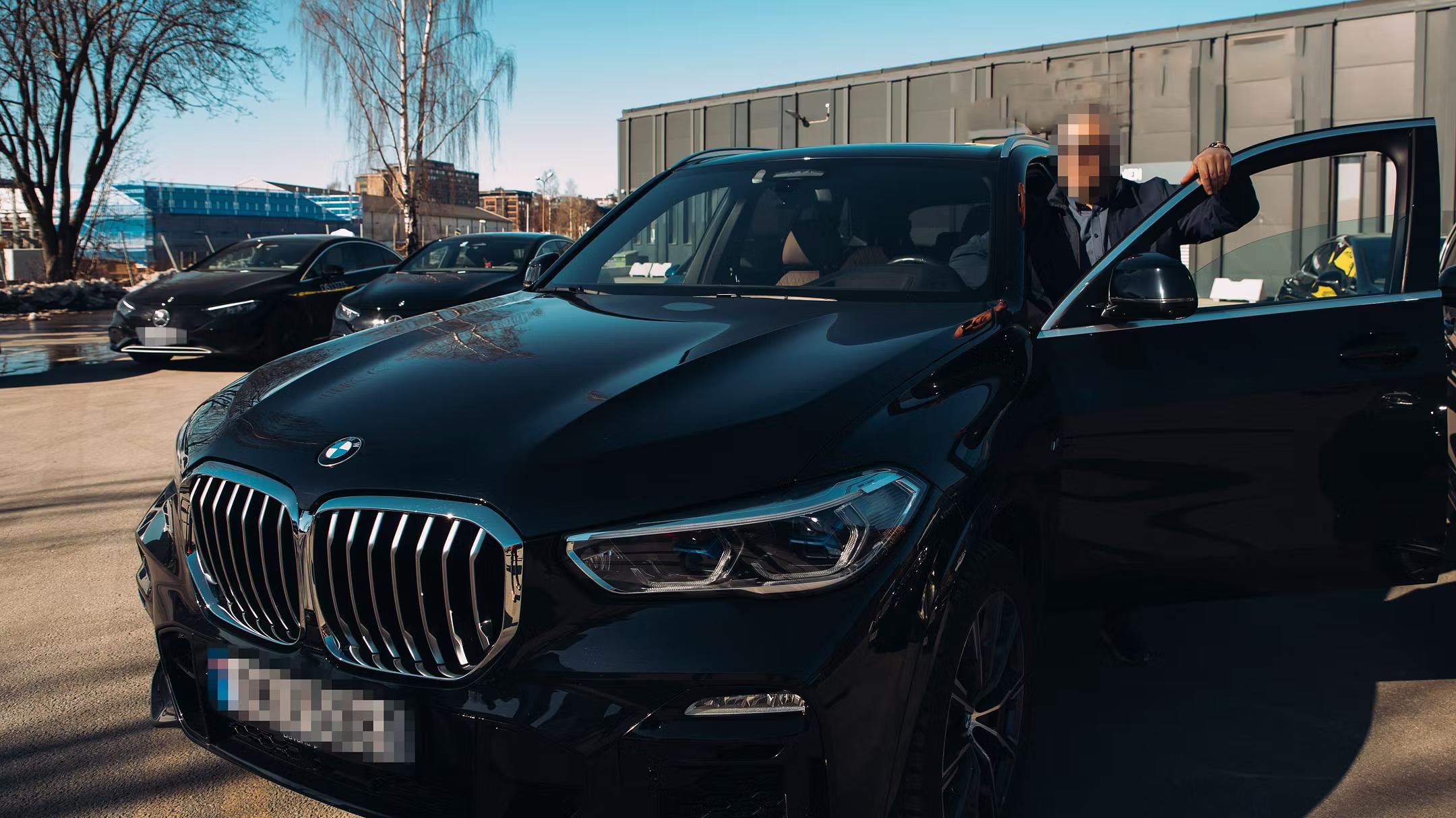 कालो BMW गाडीको चालकतर्फको ढोका खोलेर छेउमा उभिएको एक व्यक्ति। पृष्ठभूमिमा अन्य गाडी र भवन देखिन्छन्।