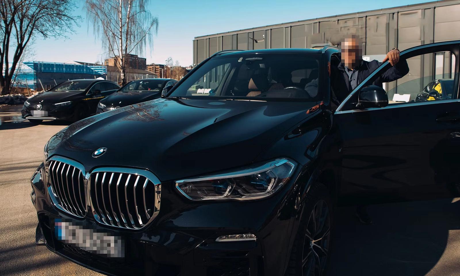 कालो BMW गाडीको चालकतर्फको ढोका खोलेर छेउमा उभिएको एक व्यक्ति। पृष्ठभूमिमा अन्य गाडी र भवन देखिन्छन्।