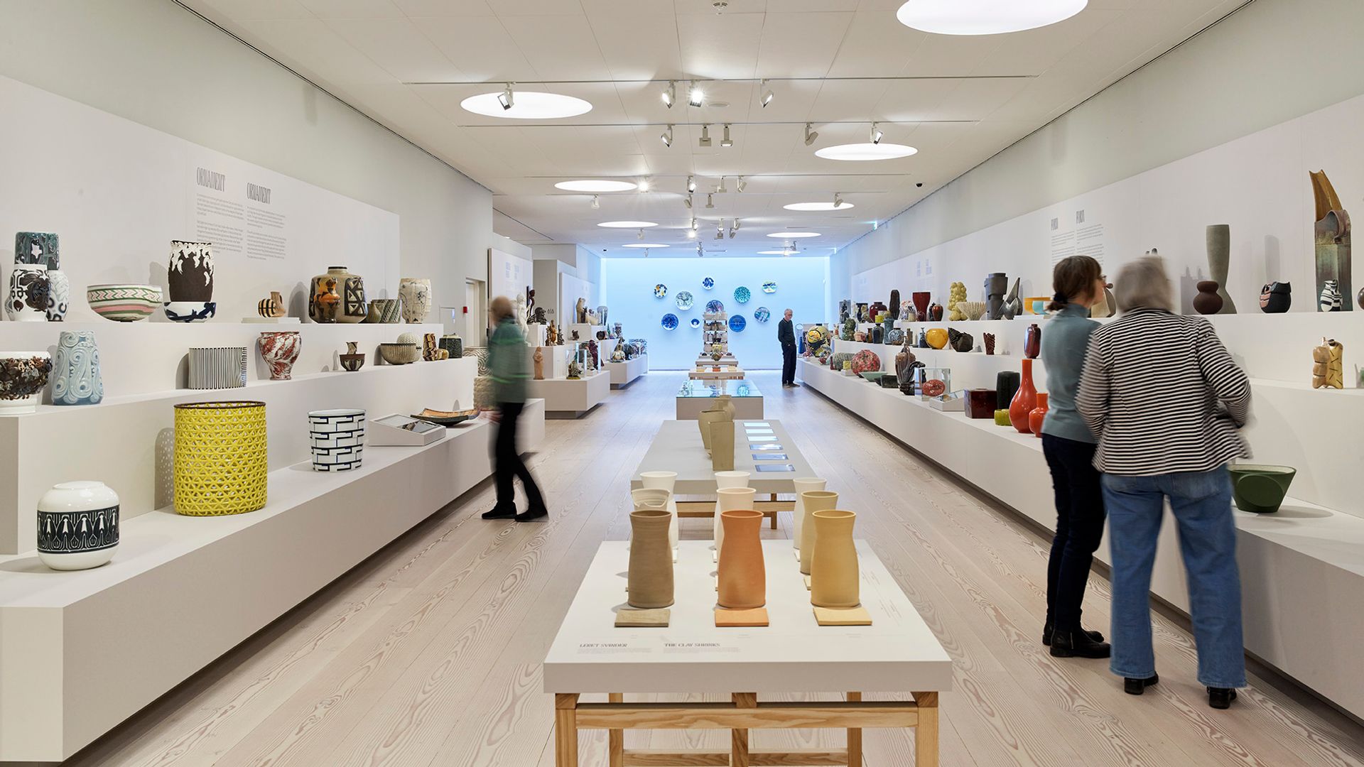 udstilling på Clay museum. Tre mennesker kigger på keramik. Der er et bord i midten og to "trapper" i siden med keramik på
