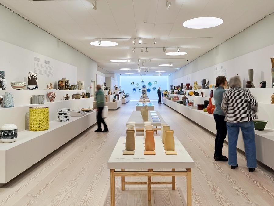 udstilling på Clay museum. Tre mennesker kigger på keramik. Der er et bord i midten og to "trapper" i siden med keramik på