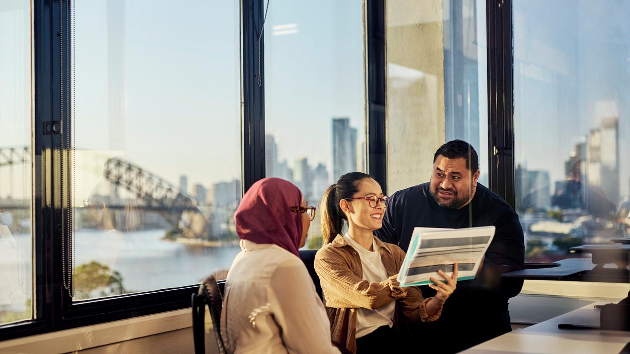 Three colleagues look over a printed report by an office window overlooking the Sydney Harbour Bridge