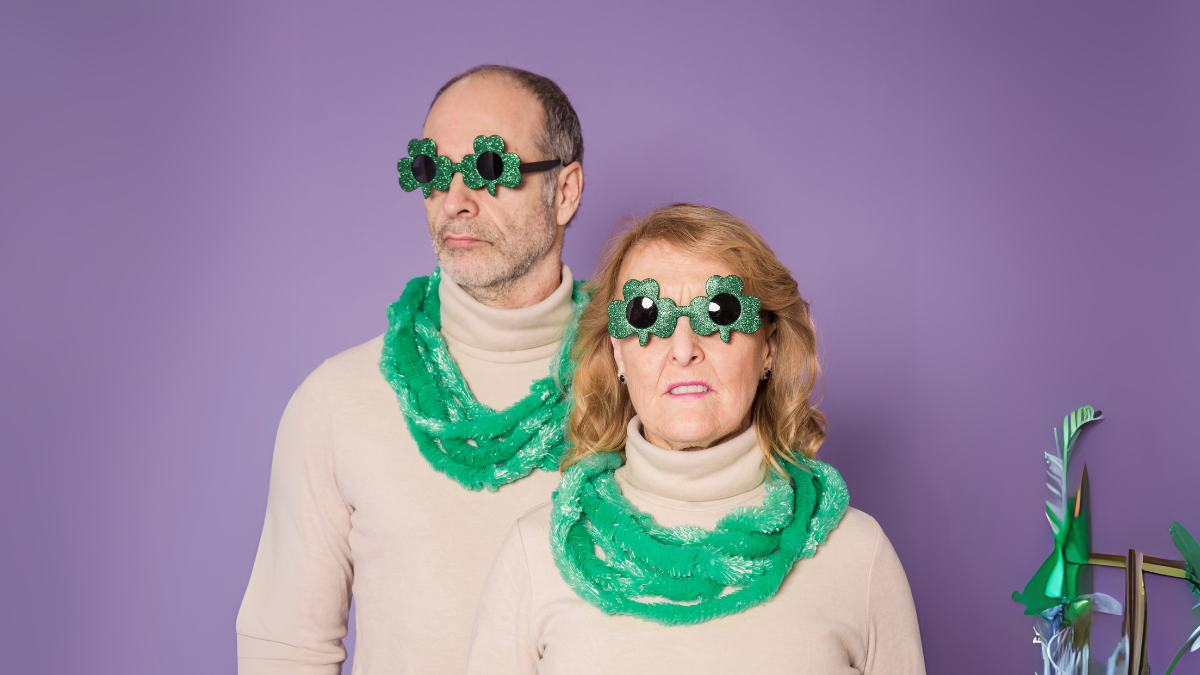 Una pareja con cuellos altos beige idénticos, bufandas verdes y gafas festivas en forma de trébol, posando contra un fondo morado