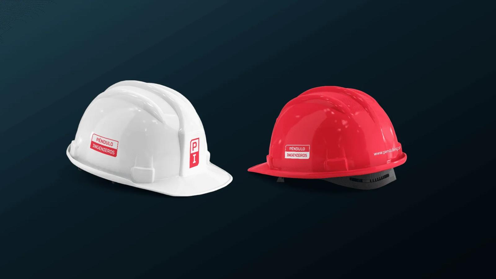 Dos cascos de seguridad industrial, uno blanco y otro rojo, colocados sobre un fondo oscuro y personalizados con el logotipo de 'Péndulo Ingenieros' y el isotipo 'PI' en los laterales