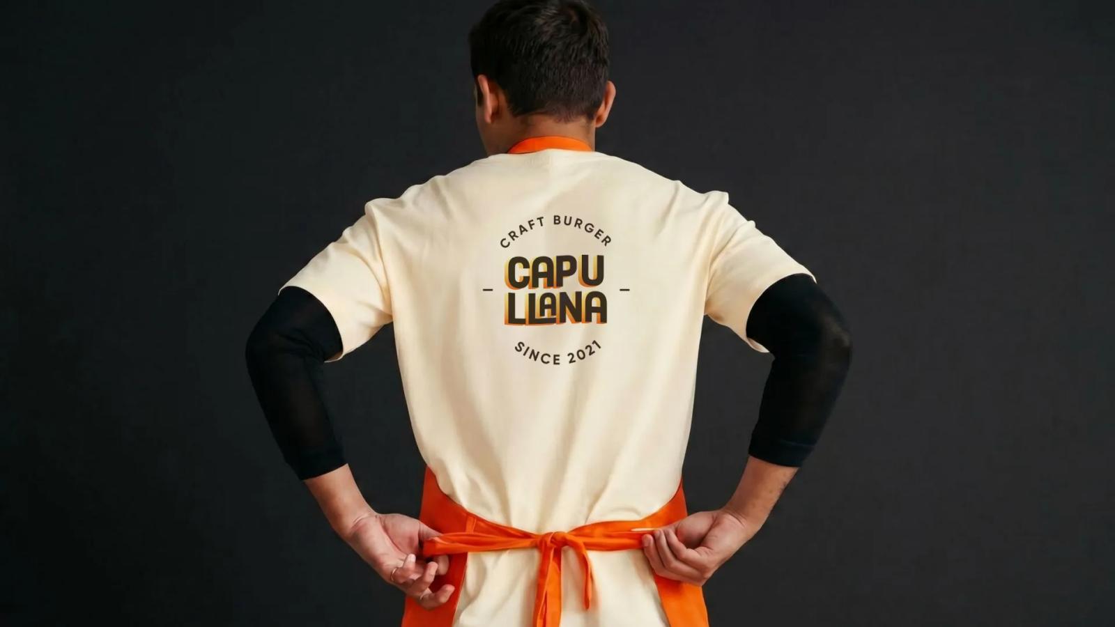 Persona de espaldas con camiseta clara de Capullana Craft Burger atándose un mandil naranja sobre fondo negro