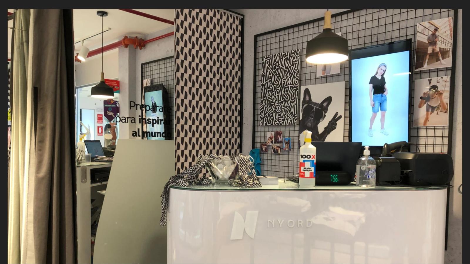 Interior de tienda física Nyord mostrando mostrador blanco con logo pared de rejilla negra con fotografías de campaña y pantalla digital exhibiendo outfits en un espacio de diseño industrial moderno