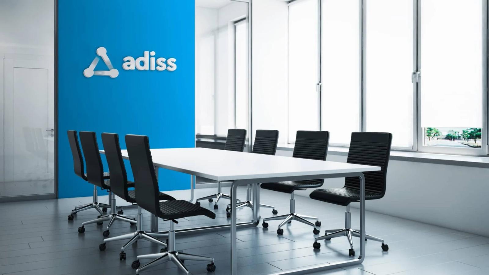 Interior de una sala de reuniones moderna con una mesa blanca rectangular rodeada de sillas negras, presentando una pared azul brillante al fondo con el logotipo de "adiss" en relieve blanco y grandes ventanales a la derecha
