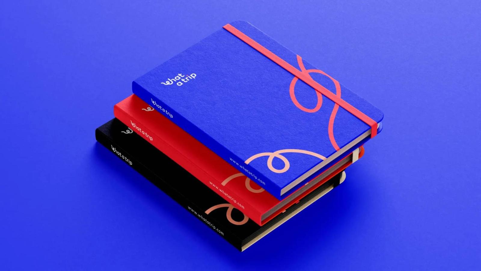 Tres cuadernos de notas corporativos de What a Trip apilados en colores azul rojo y negro con el logotipo y diseño de líneas onduladas sobre fondo azul eléctrico