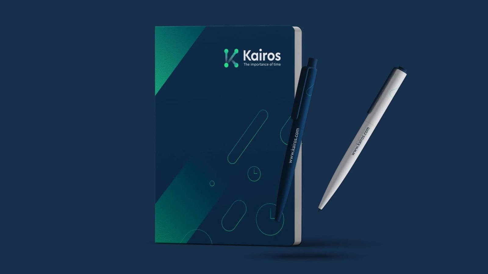 Libreta azul oscuro con diseño geométrico y logotipo junto a dos bolígrafos promocionales en azul y blanco con la dirección web impresa sobre fondo oscuro y lapicero al lado derecho