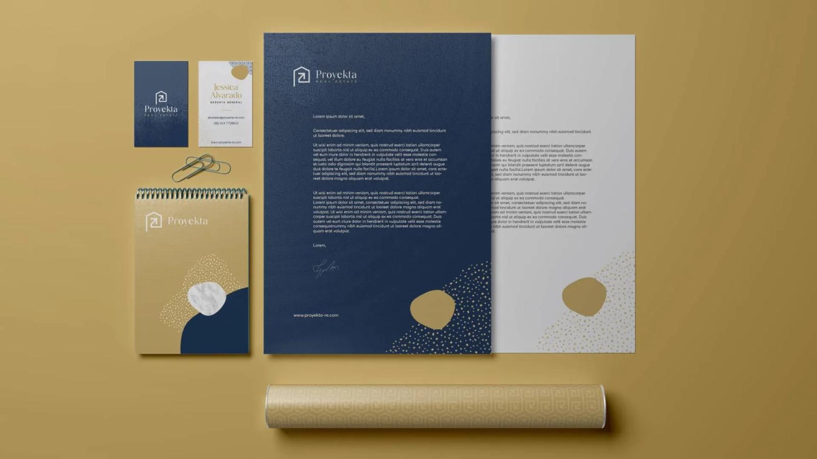 Papelería corporativa Proyekta incluyendo tarjetas de presentación libreta dorada hoja membreteada azul y tubo de planos con diseño de marca inmobiliaria sobre fondo mostaza