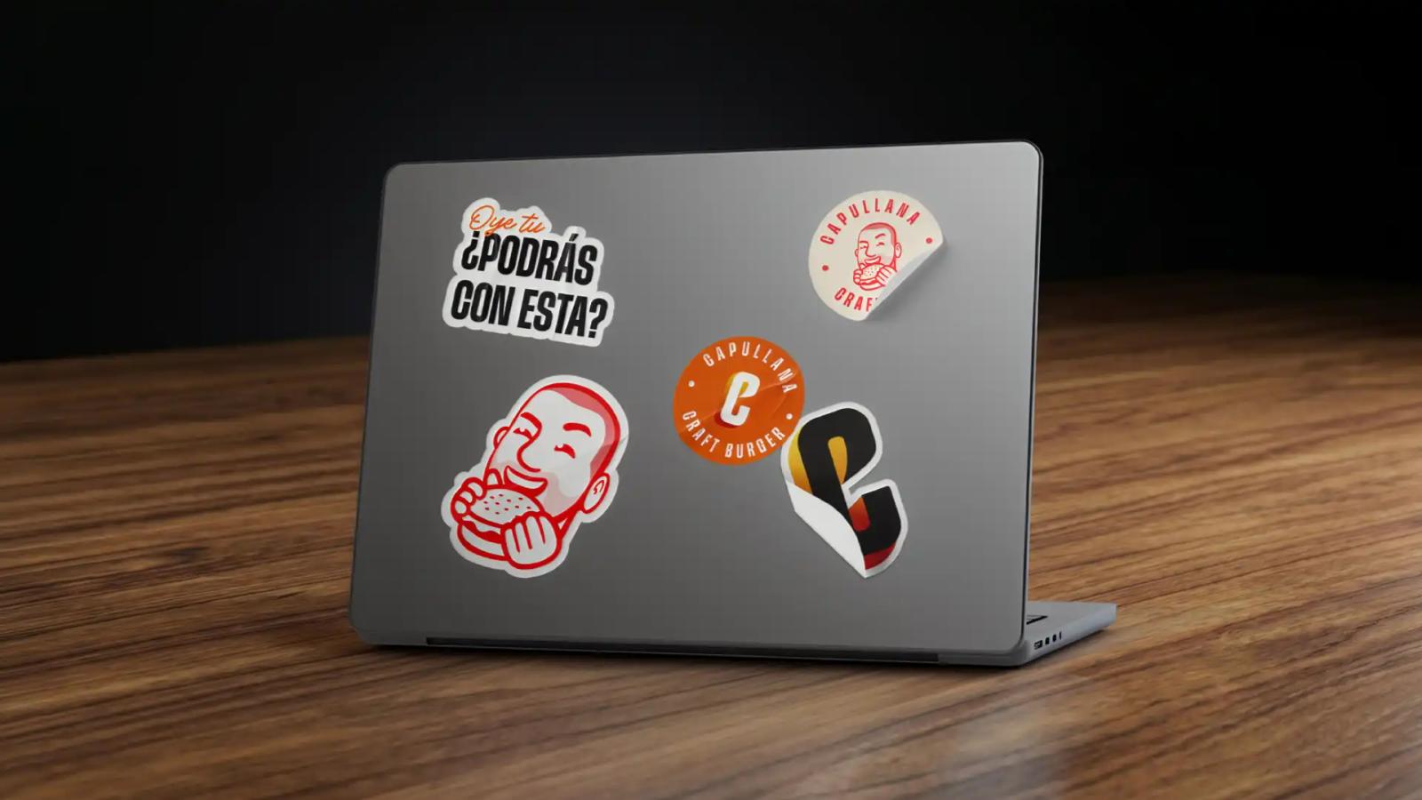 Laptop gris sobre una mesa de madera con varios stickers de la marca Capullana Craft Burger pegados en la tapa