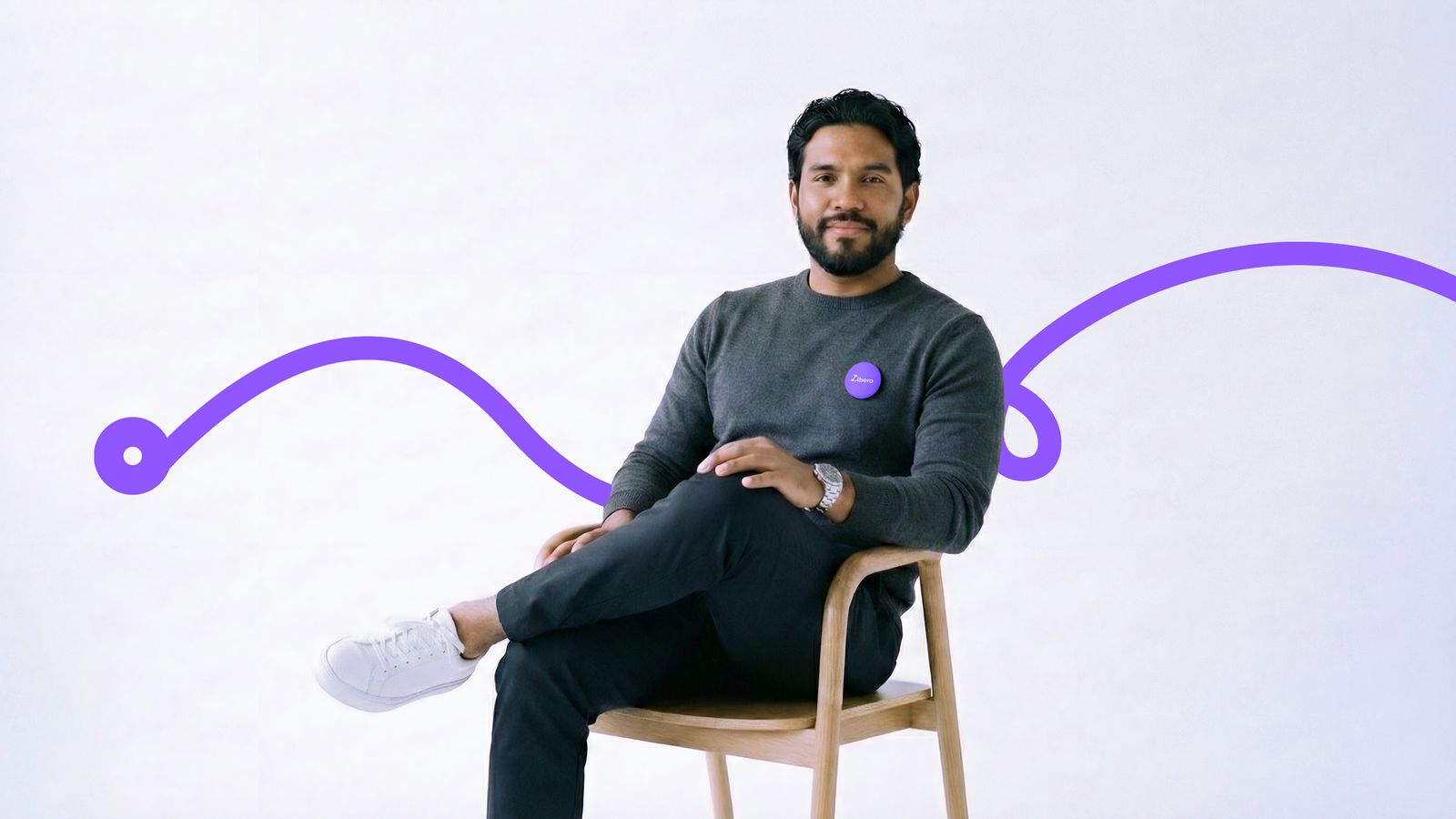 Hombre con barba sentado en una silla de madera frente a un fondo blanco, decorado con una línea ondulada violeta y portando un pequeño pin morado en su suéter gris