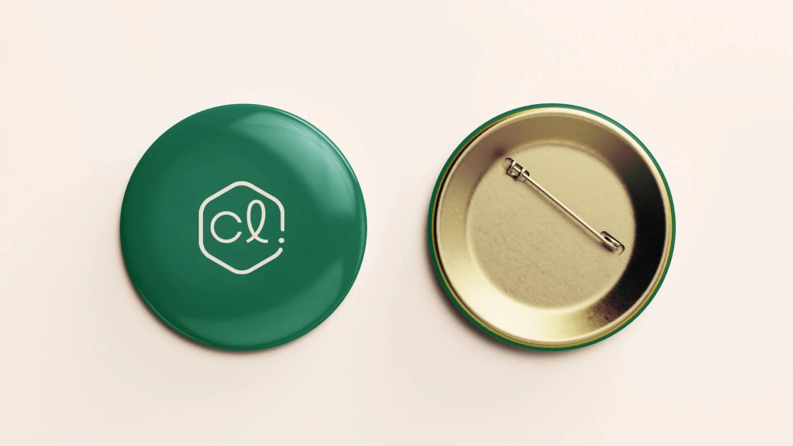 Dos botones circulares tipo chapa sobre fondo beige: a la izquierda, el anverso verde oscuro brillante con el logotipo en un hexágono blanco; a la derecha, el reverso metálico dorado mostrando el alfiler de sujeción