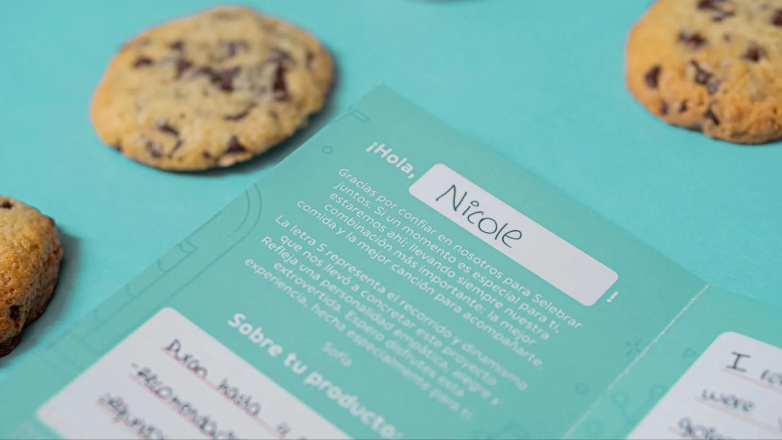 Primer plano cenital de una tarjeta de agradecimiento color turquesa personalizada, rodeada de galletas con chispas de chocolate sobre un fondo del mismo tono, explicando el significado del logotipo de la marca y detalles del producto
