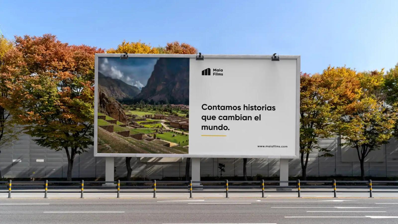 Valla publicitaria exterior de Maia Films dividida en foto de paisaje andino y eslogan Contamos historias que cambian el mundo sobre fondo blanco entre árboles de follaje otoñal y cielo azul despejado