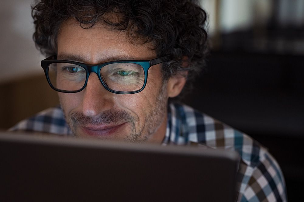 Nahaufnahme eines Mannes mit lockigem Haar und Brille, der konzentriert auf den Bildschirm eines Laptops blickt. Er trägt ein kariertes Hemd und lächelt leicht.