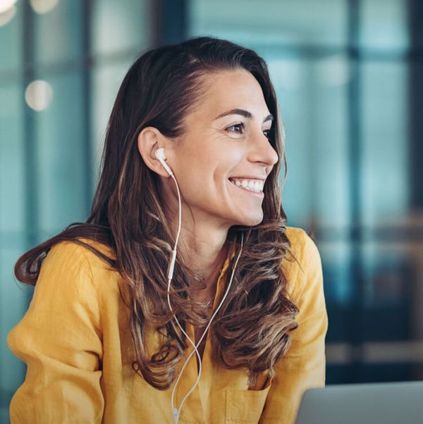 Engagierte, junge Dame, gelber Pulli mit In-Ear-Kopfhörern lächelt erfreut, sitzend vor Ihrem Notebook in einem modernen Büroraum.
