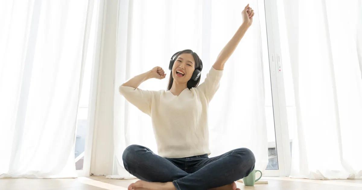 หญิงสาวใส่หูฟังนั่งขัดสมาธิบนพื้นห้องที่มีแสงแดดส่อง ยกแขนยิ้มอย่างผ่อนคลาย สื่อถึงการจัดการความเครียดและอารมณ์ที่ดี