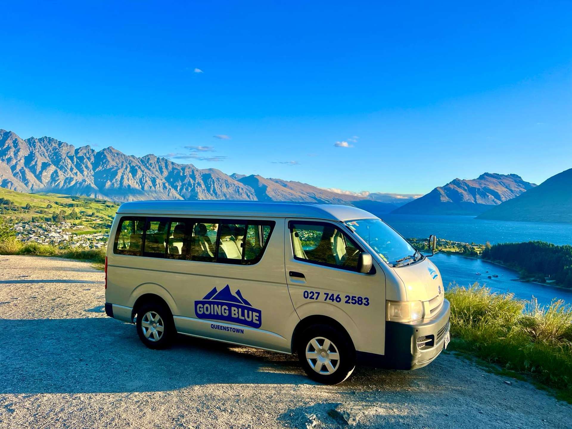 Going Blue Adventures tour van parked at a scenic viewpoint above Queenstown with Lake Wakatipu and The Remarkables mountain range in the background.
