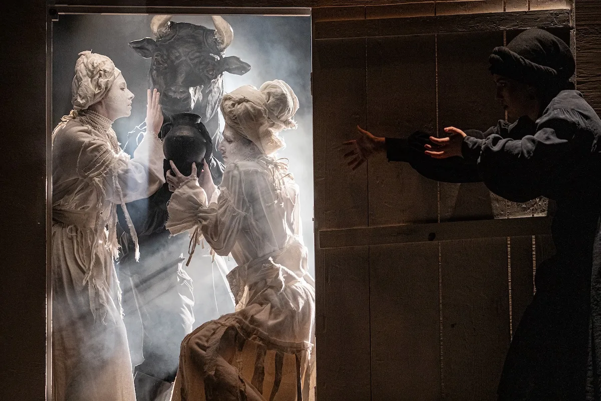 Performers in white costumes and a bull mask in a doorway of light.