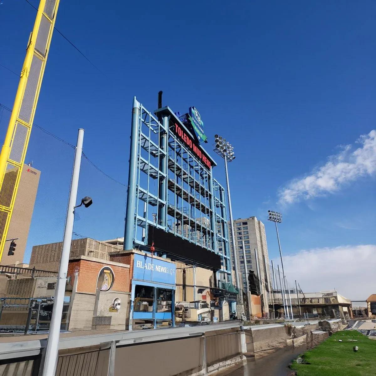 Ballpark video scoreboard and light towers behind the outfield wall under a blue sky