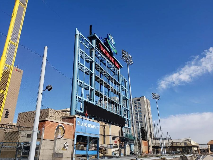 Ballpark video scoreboard and light towers behind the outfield wall under a blue sky