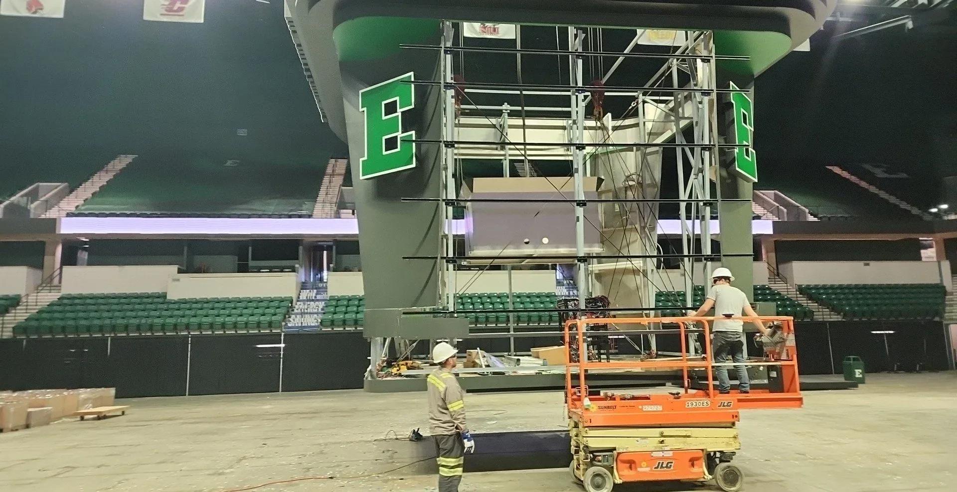 Scissor lift and scaffolding under a center-hung scoreboard frame in an arena with green E logos
