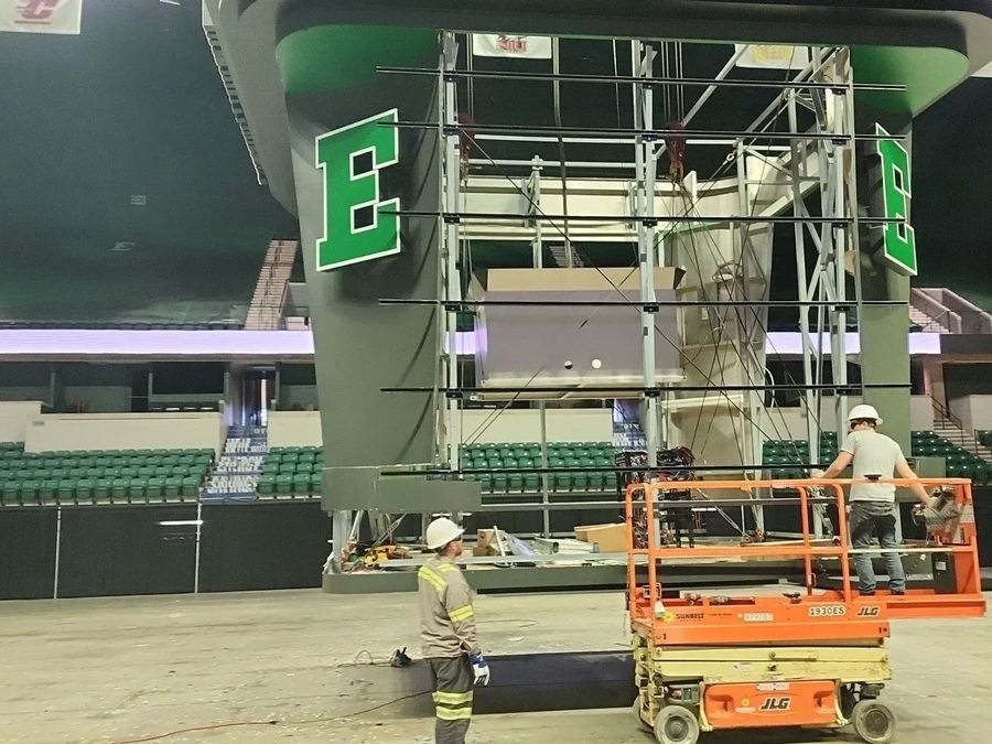 Scissor lift and scaffolding under a center-hung scoreboard frame in an arena with green E logos