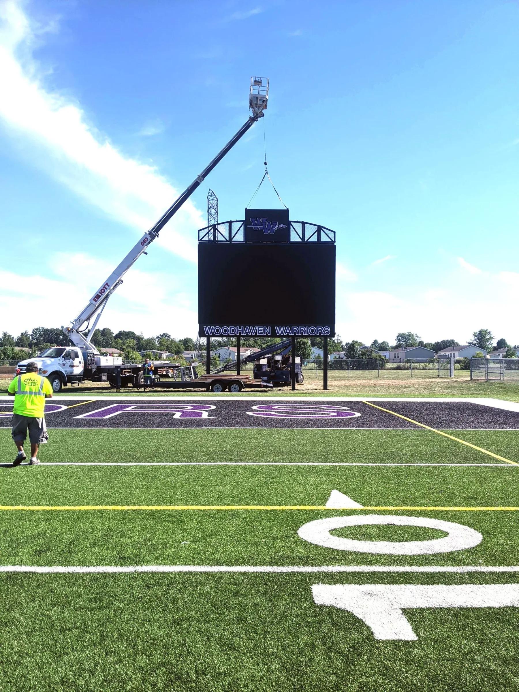 Crane setting a scoreboard beside a football field while an installer in a safety vest watches