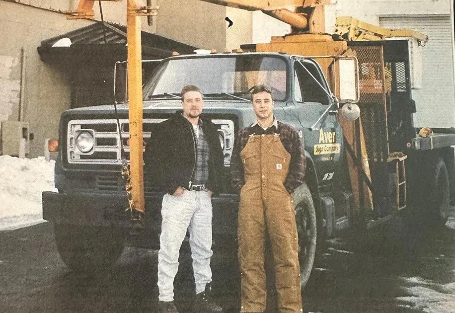 Archive photo of two young men standing beside an early Aver Sign Company boom truck in the snow