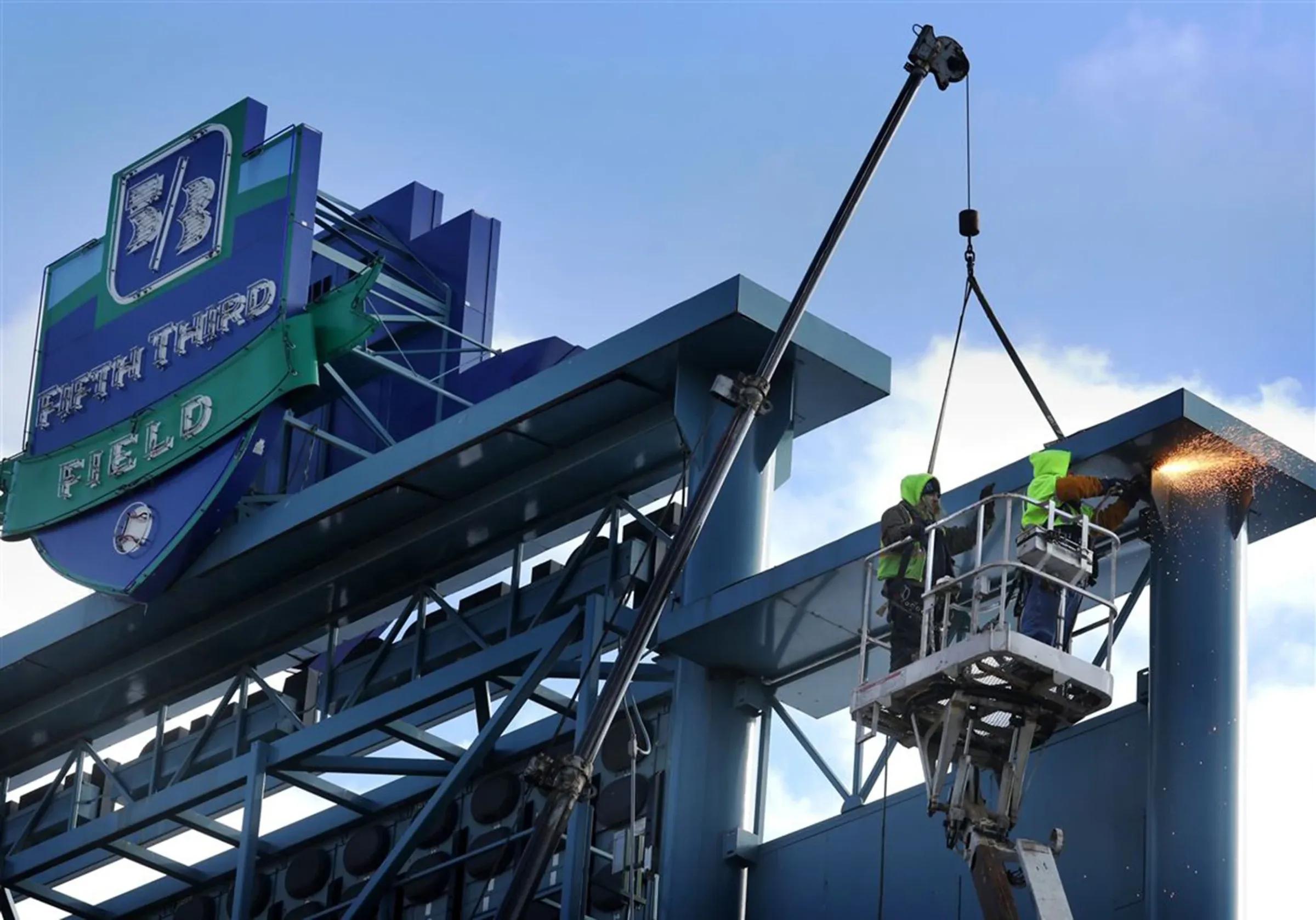 Crane holding a steel section while two workers in a lift weld beside the Fifth Third Field sign
