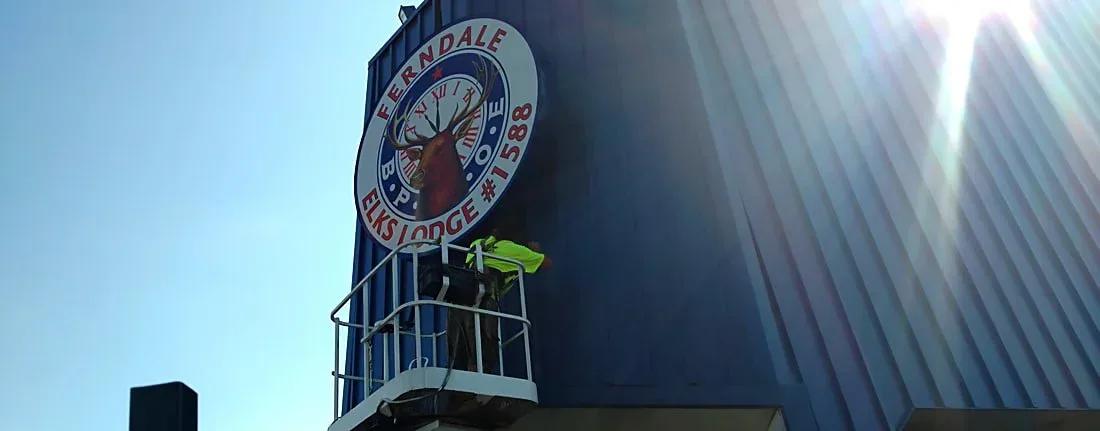 Installer in a lift beside a round Ferndale Elks Lodge sign on a building corner