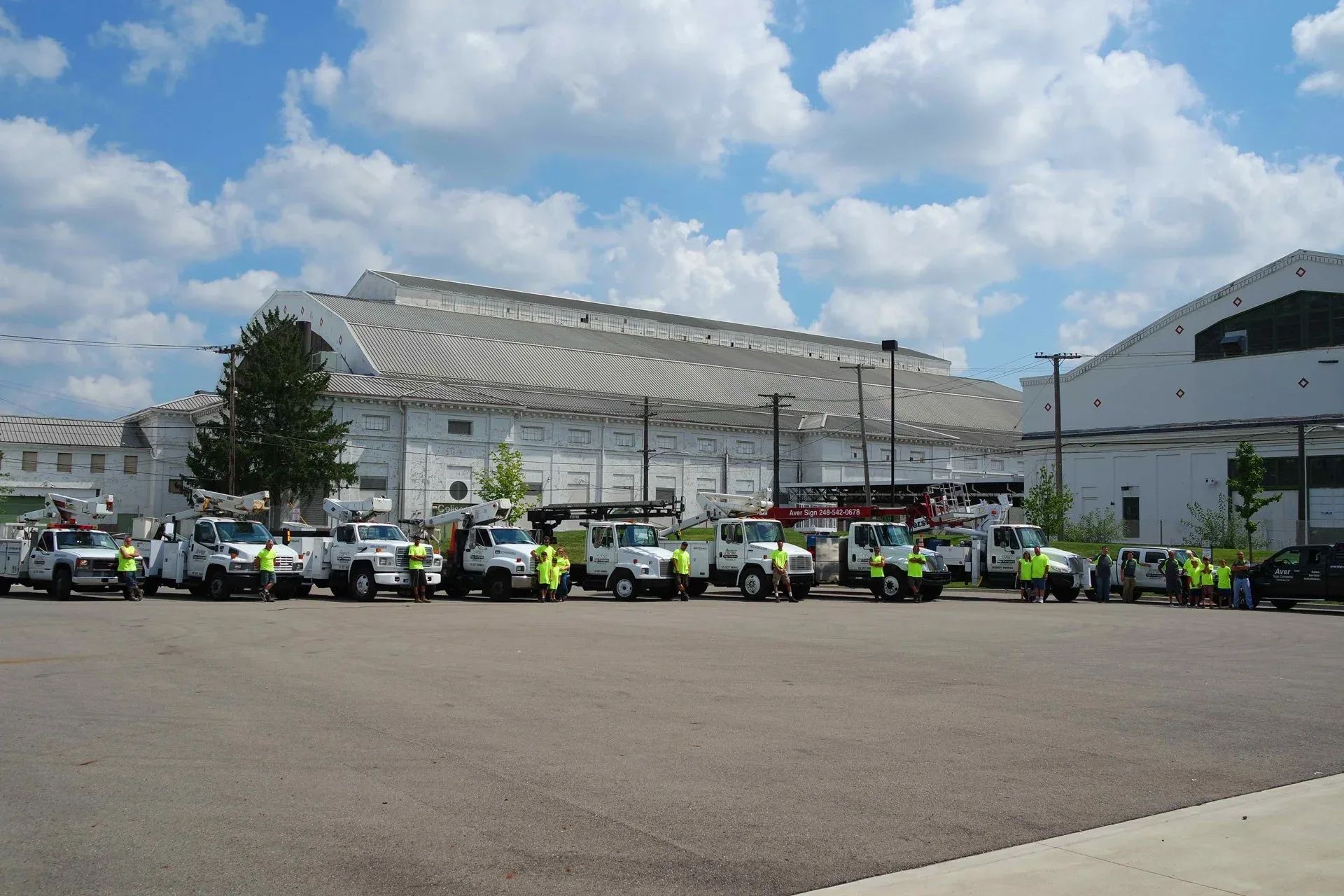 Row of Aver Sign Company bucket and crane trucks with crew members in front of a large white building