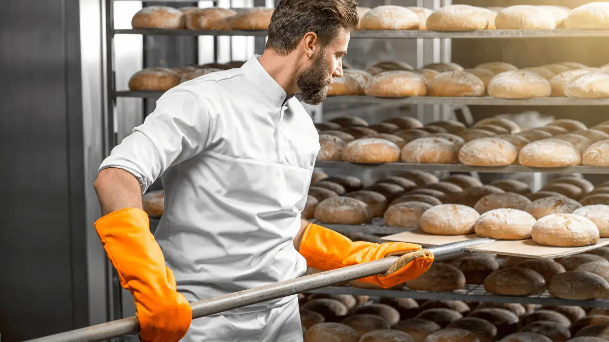 Een bakker in een witte bakkersjas en feloranje hittebestendige handschoenen gebruikt een houten schep om ronde broden te plaatsen of te verwijderen van een grote metalen rek vol met versgebakken brood in een industriële bakkerijomgeving.