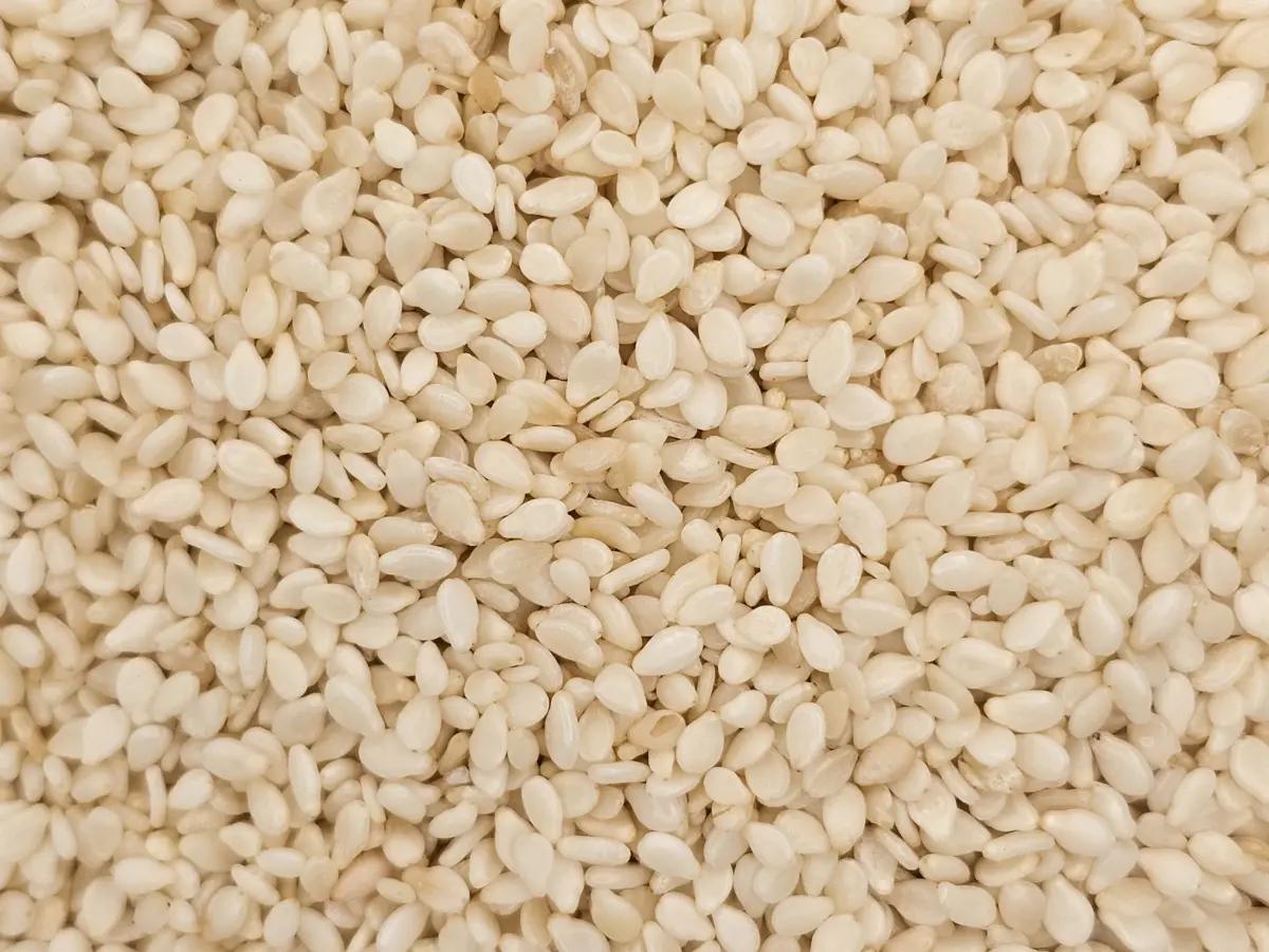 Close-up view of a large pile of hulled sesame seeds, small and oval-shaped, off-white in color, densely packed together.